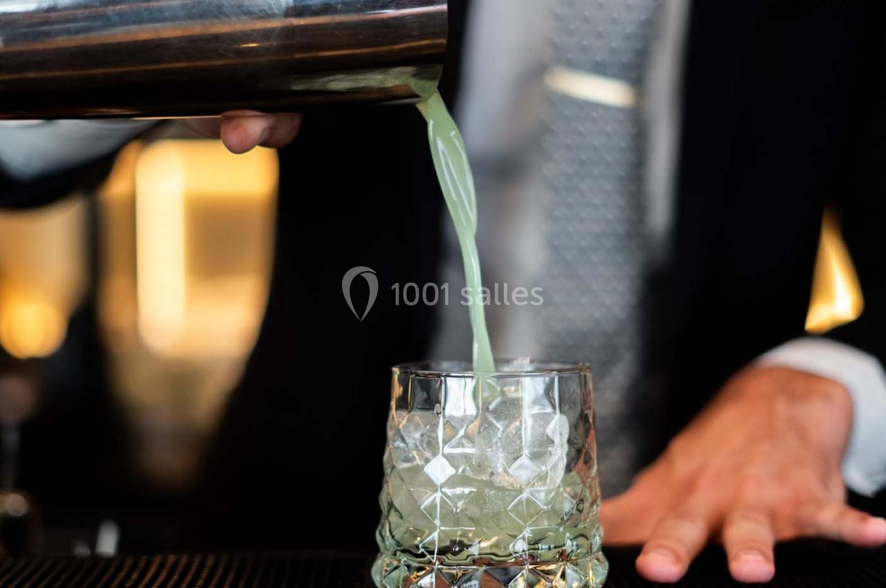 Un barman verse un cocktail vert dans un verre en cristal sur un comptoir.