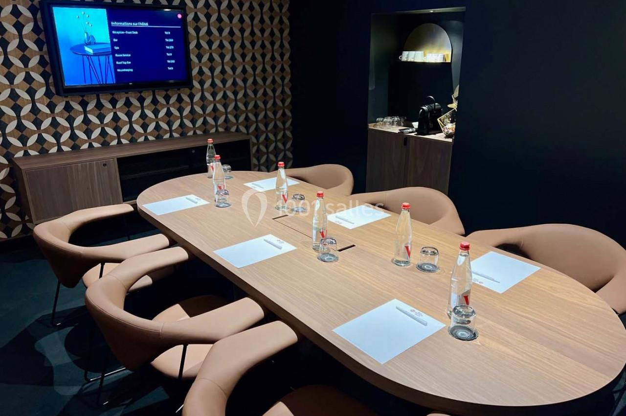 Salle de réunion moderne avec table ovale, chaises en cuir, bouteilles d'eau, blocs-notes et écran mural allumé.