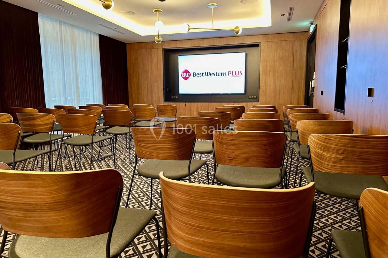 Salle de conférence avec rangées de chaises en bois, écran de présentation allumé et éclairage suspendu.