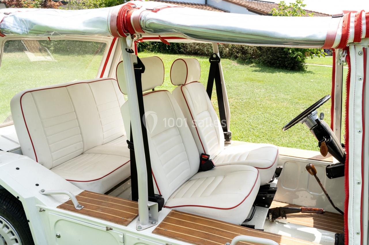 Intérieur d'une voiture décapotable vintage avec sièges blancs bordés de rouge, vue sur un jardin en arrière-plan.