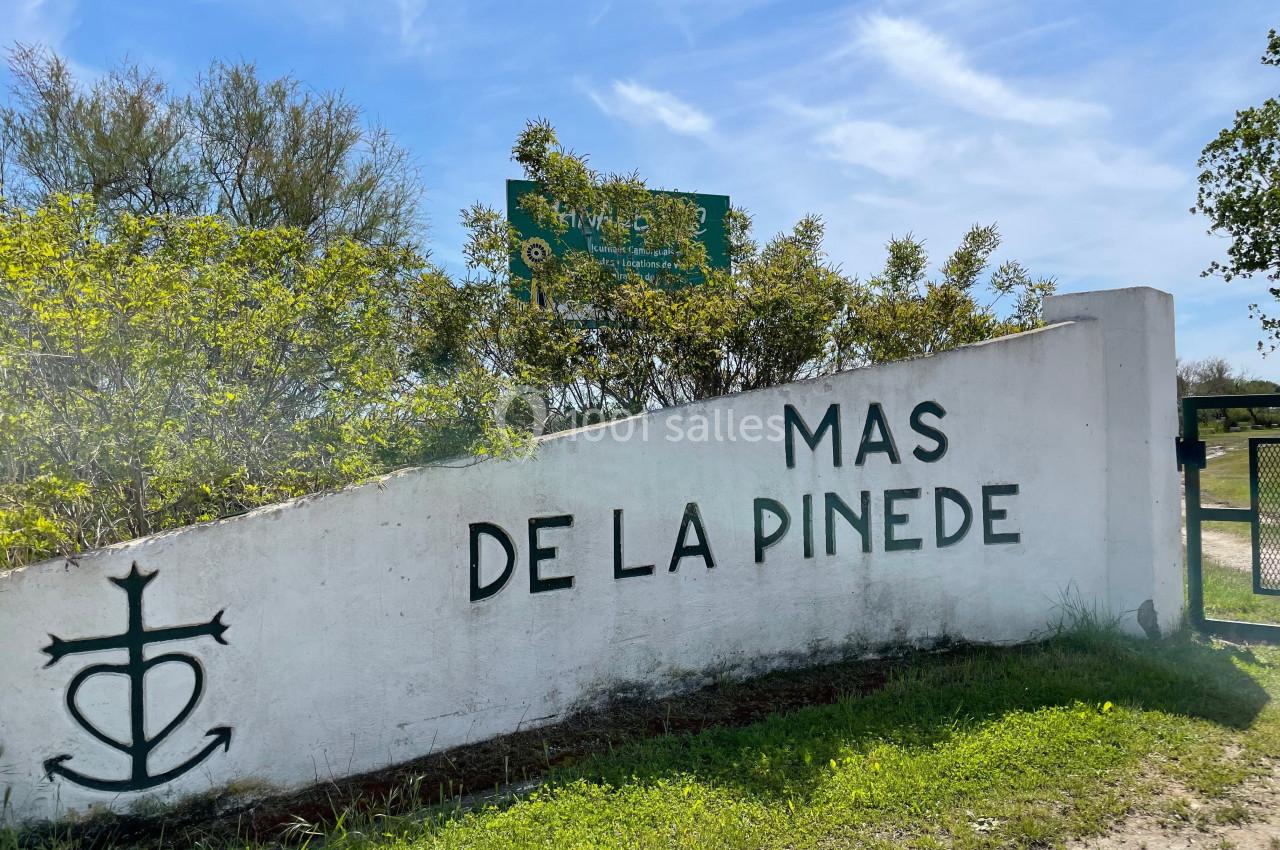 Mur blanc avec l'inscription ’Mas de la Pinède’ et une croix camarguaise, entouré de végétation sous un ciel bleu.