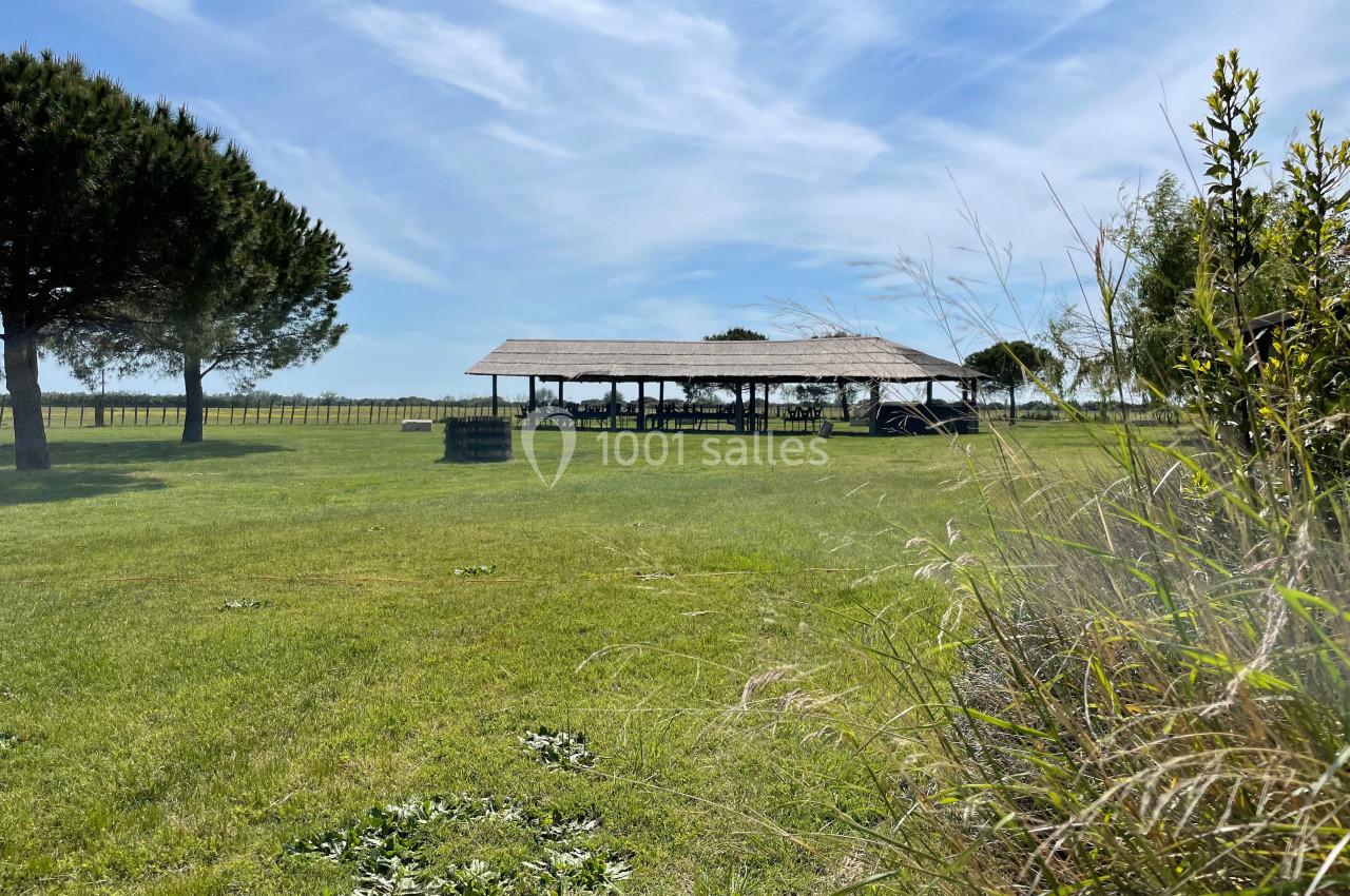 Abri en bois au milieu d'un pré verdoyant, entouré d'arbres et de végétation sous un ciel partiellement nuageux.