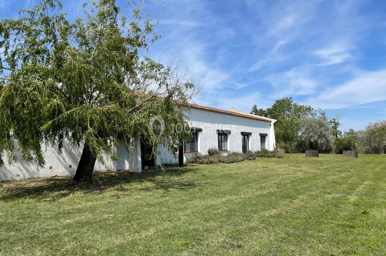 Bâtiment blanc de plain-pied avec toit en tuiles, entouré d'un grand jardin et d'arbres sous un ciel bleu.