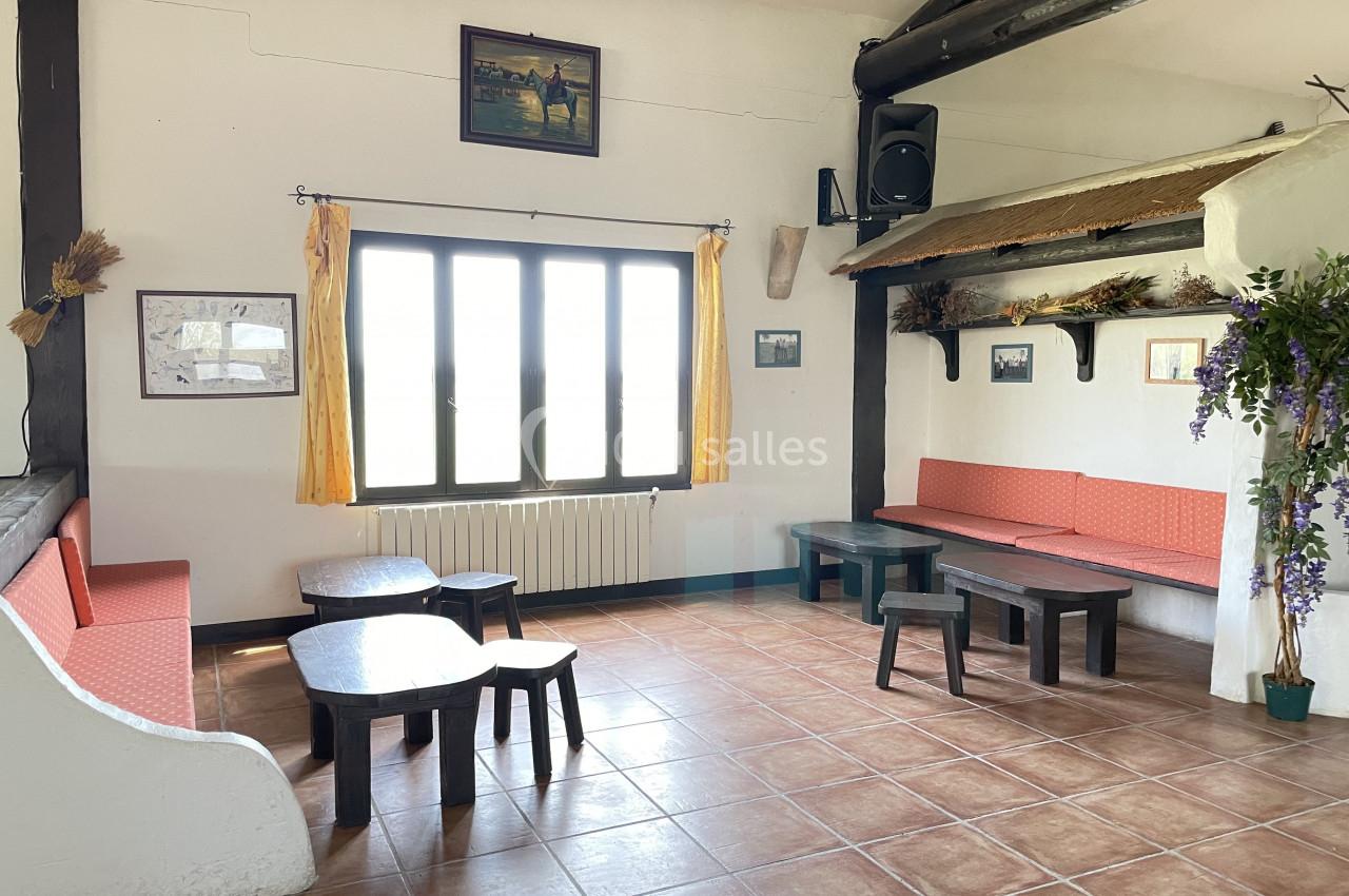 Salle lumineuse avec des bancs et tables en bois, décorée de cadres, plantes et rideaux jaunes devant une grande fenêtre.