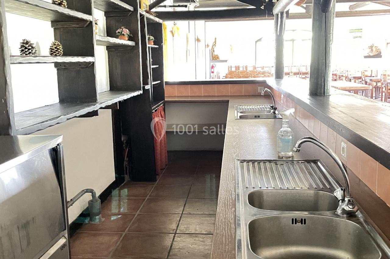 Cuisine ouverte avec plan de travail en bois, double évier en inox, étagères murales et carrelage au sol.