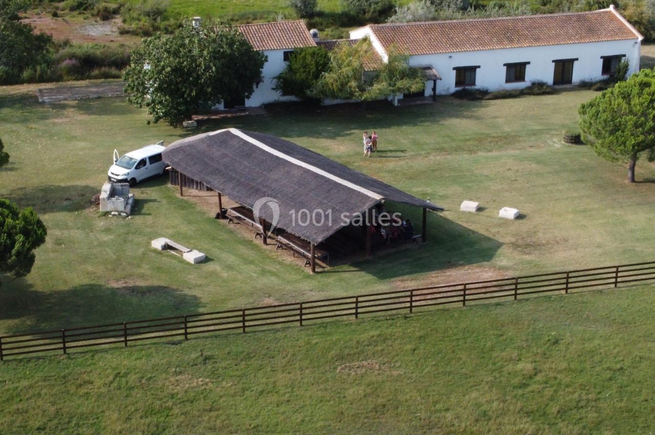 Vue aérienne d'une structure en bois avec toit en chaume dans un espace vert, entourée d'arbres et de bâtiments.