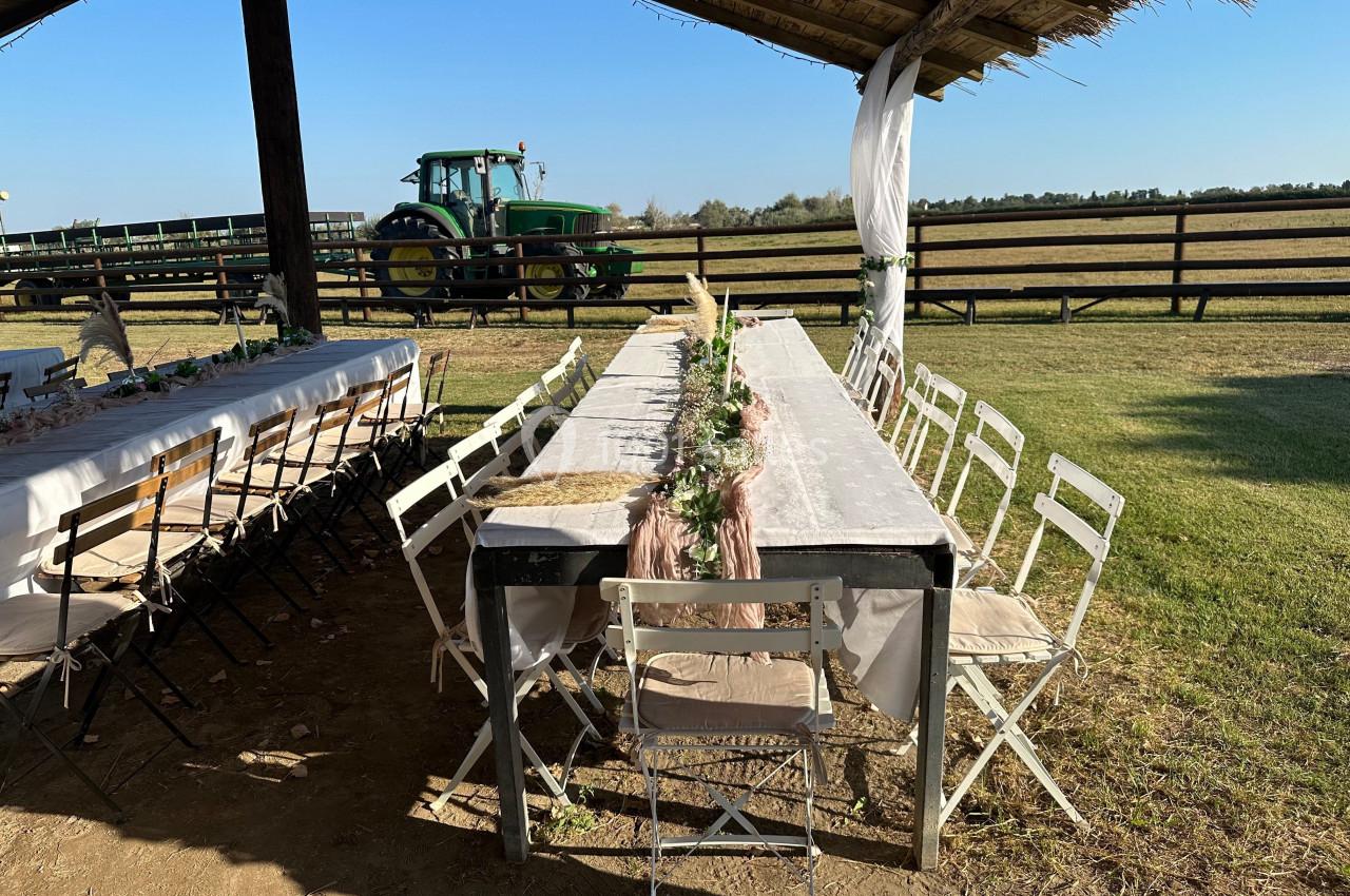 Tables décorées pour un repas en extérieur sous un abri en bois, avec un tracteur et des champs en arrière-plan.