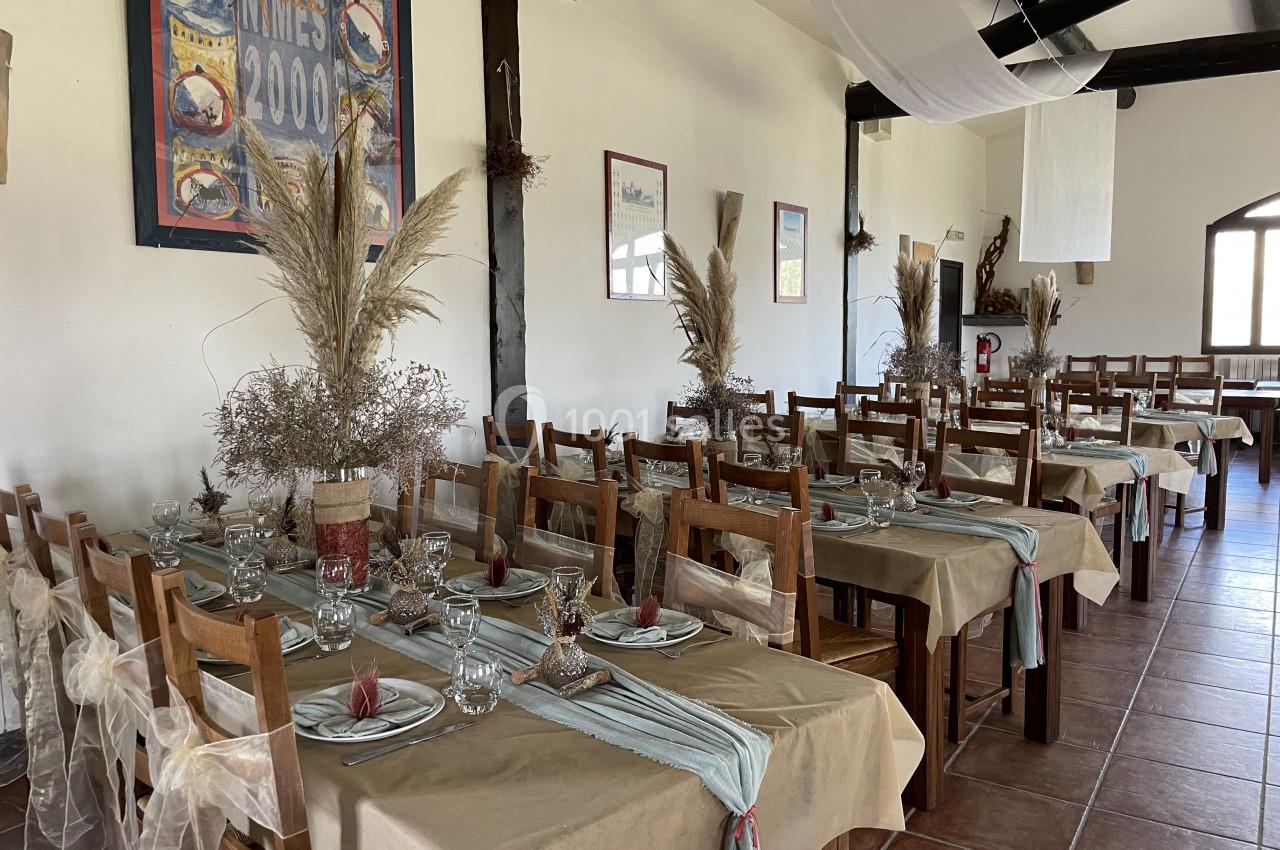 Salle de réception décorée avec des tables alignées, nappes beiges, chemins de table bleus et bouquets de fleurs séchées.