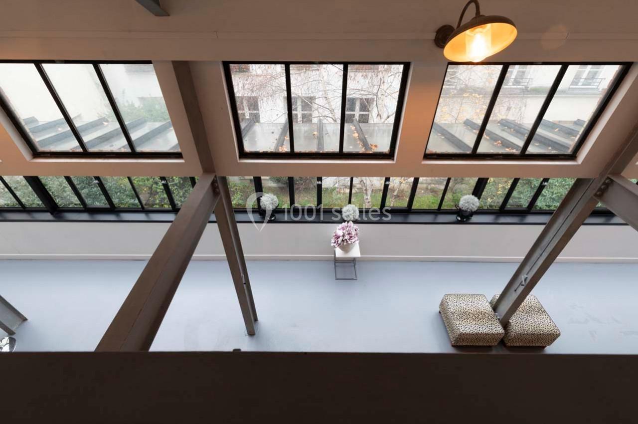 Vue intérieure d'un espace lumineux avec de grandes fenêtres, un sol gris, des tabourets et une table décorée de fleurs.