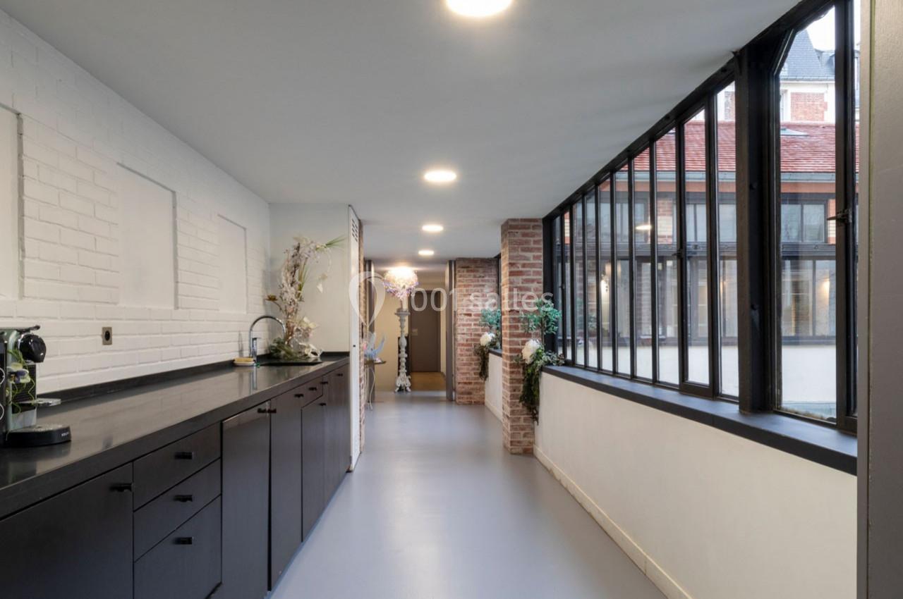 Cuisine lumineuse avec plan de travail noir, mur en briques blanches et grandes fenêtres donnant sur une cour.