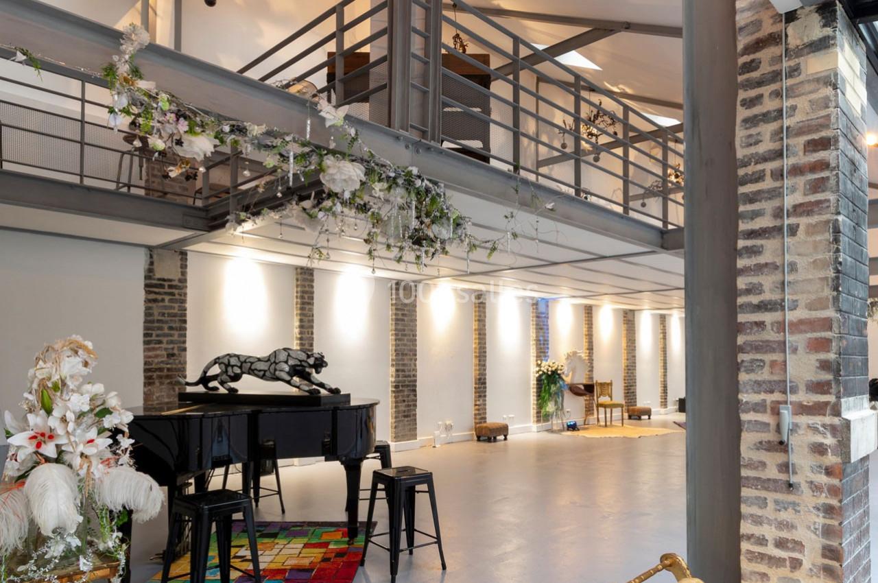 Intérieur spacieux avec mezzanine, piano noir, tabourets, décor floral et murs en briques apparentes.