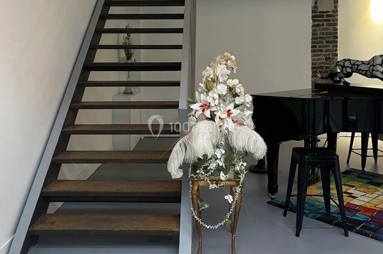 Escalier en bois à côté d'une composition florale blanche, avec un piano et un tapis coloré en arrière-plan.