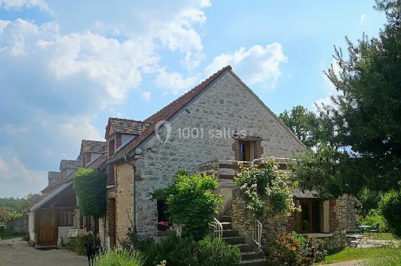 Maison en pierre entourée de verdure, avec un ciel bleu et quelques nuages en arrière-plan.