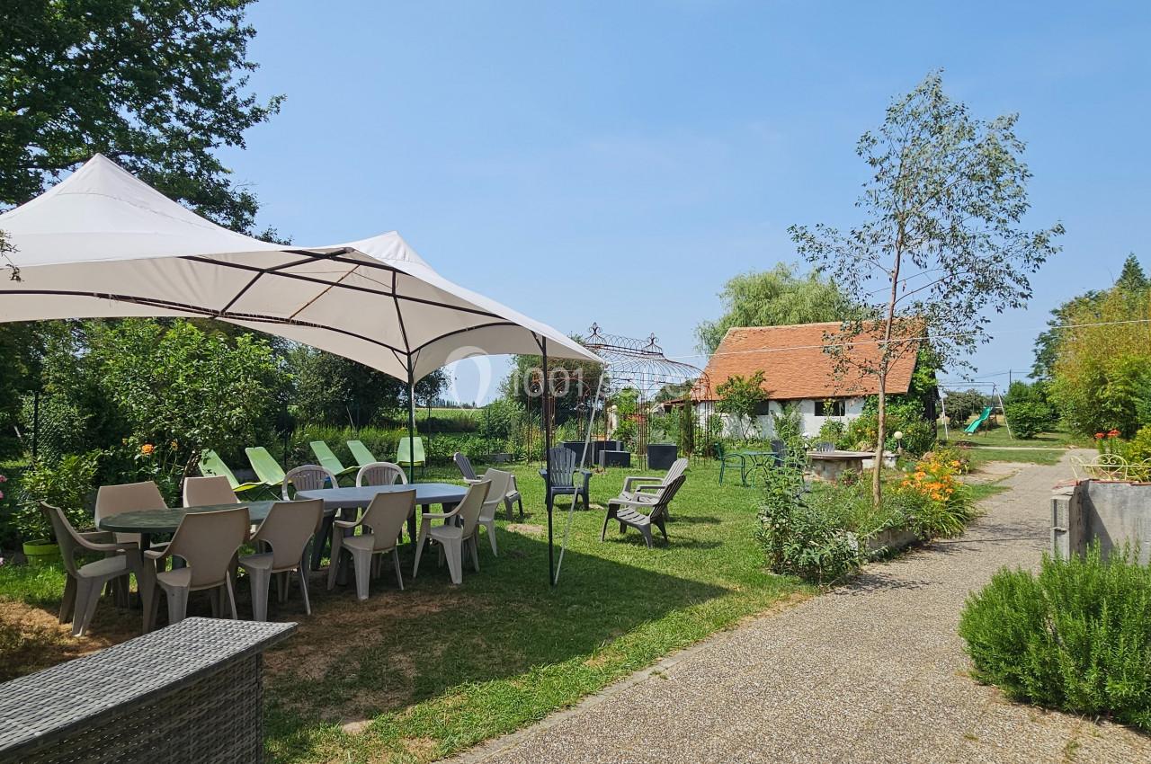 Jardin avec tables et chaises sous un grand parasol blanc, entouré de verdure et de bâtiments en briques.