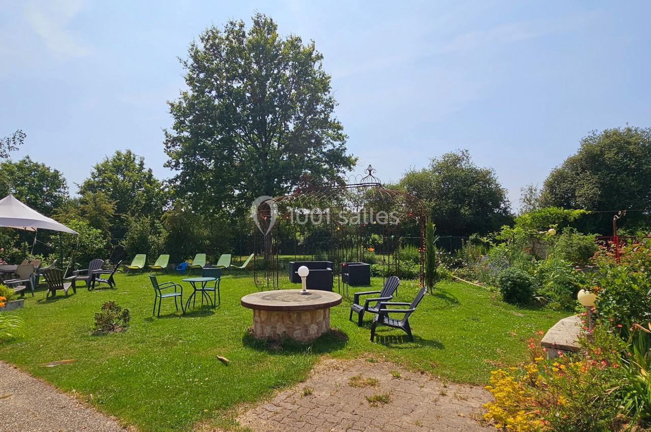 Espace extérieur avec pelouse, chaises, table ronde en pierre, parasol et végétation environnante sous un ciel dégagé.
