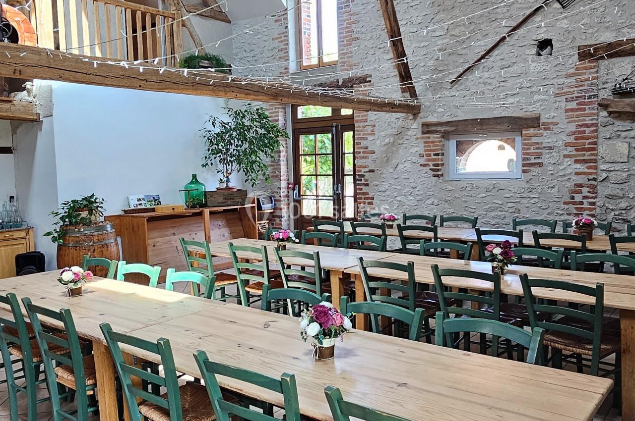 Salle lumineuse avec de grandes tables en bois, chaises vertes, poutres apparentes et murs en pierre.
