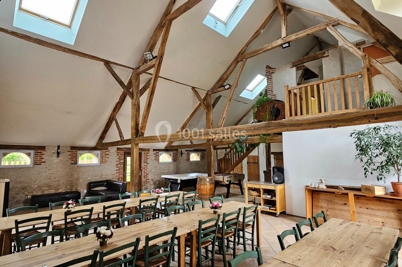 Salle lumineuse avec poutres apparentes, grandes tables en bois, chaises vertes et baies vitrées laissant entrer la lumière.