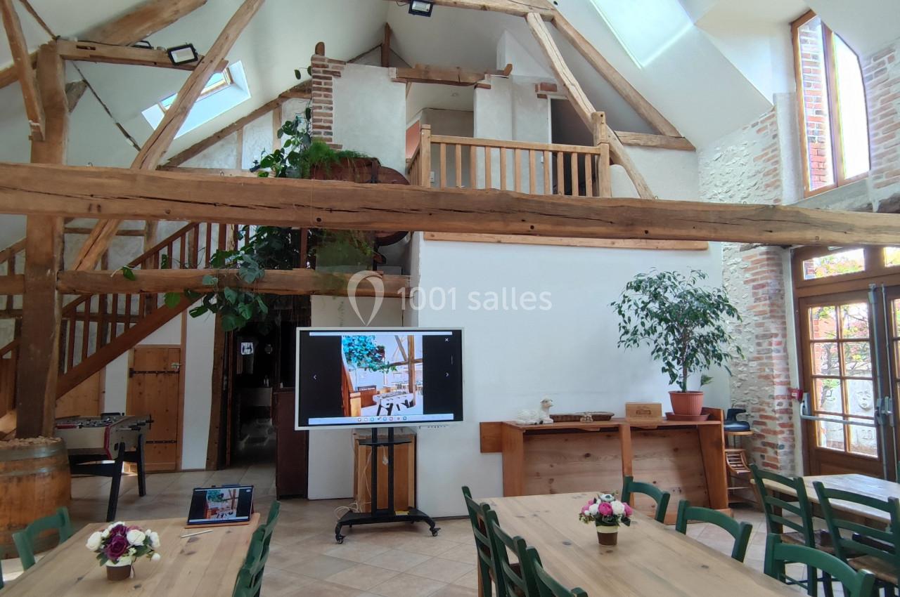 Salle lumineuse avec poutres apparentes, tables en bois, plantes, écran de présentation et billard en arrière-plan.