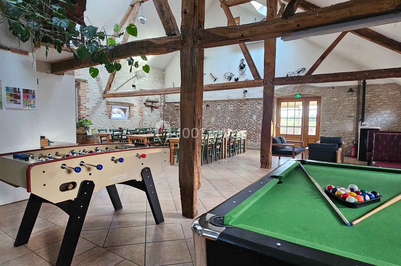 Salle conviviale avec baby-foot, table de billard, poutres apparentes et coin repas éclairé par une porte vitrée.