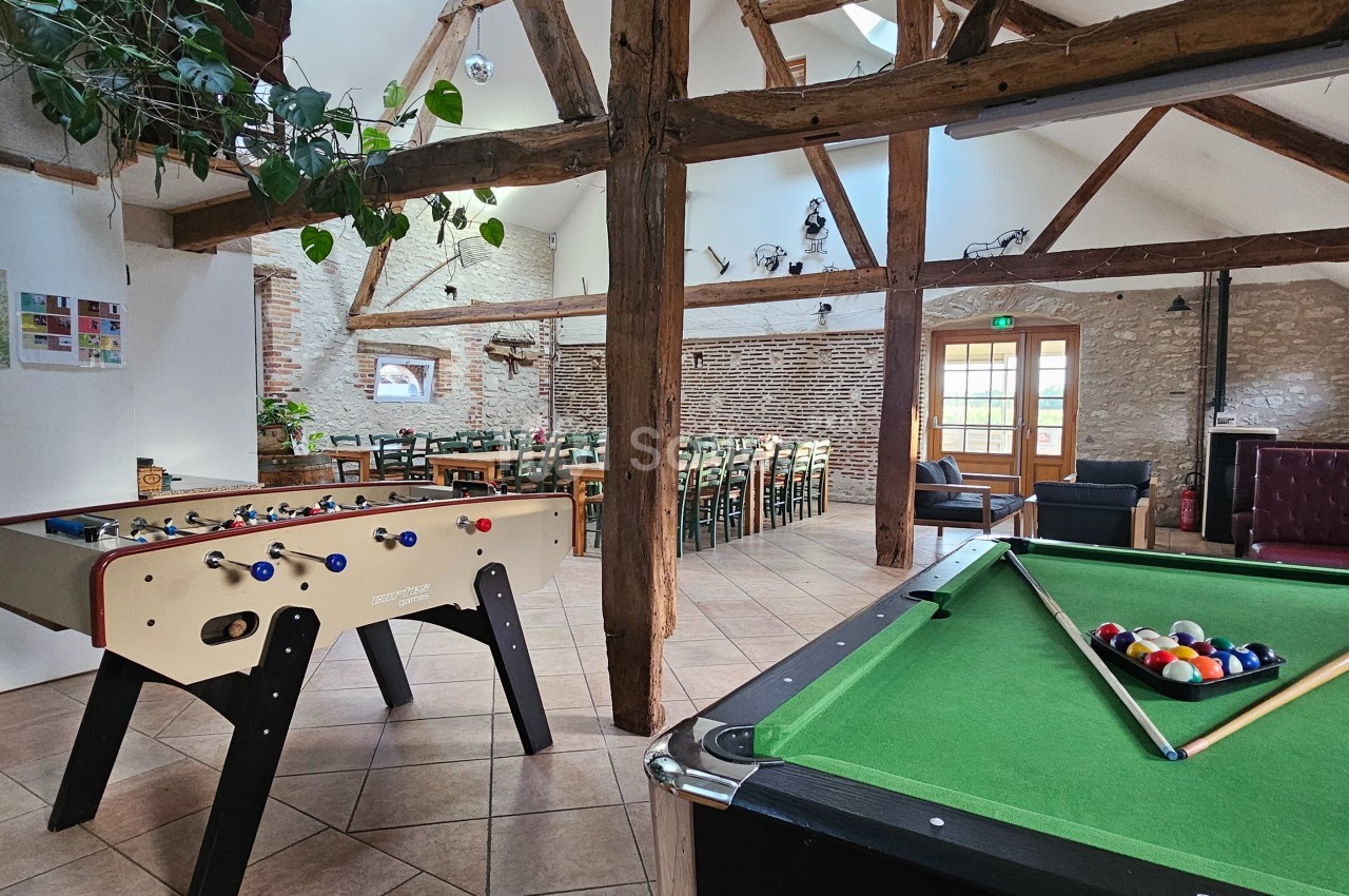Salle conviviale avec baby-foot, table de billard, poutres apparentes et coin repas éclairé par une porte vitrée.