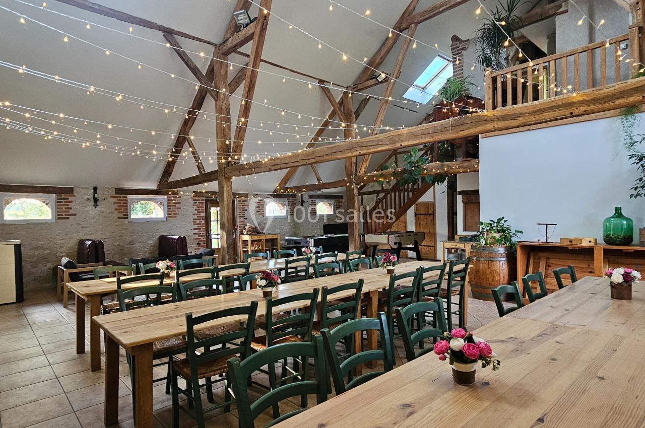 Salle lumineuse avec poutres apparentes, tables en bois, chaises vertes, guirlandes lumineuses et décorations florales.