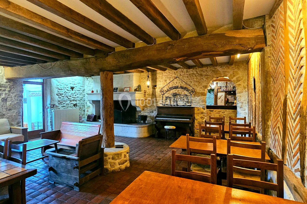 Salle rustique avec murs en pierre, poutres en bois, cheminée, piano et mobilier en bois dans un cadre chaleureux.