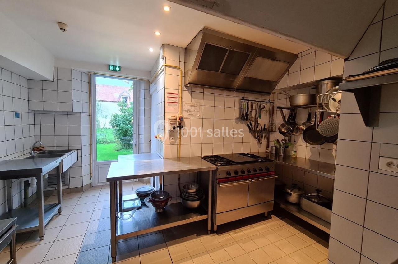 Cuisine professionnelle équipée avec plan de travail en inox, plaques de cuisson, ustensiles suspendus et vue sur un jardin.