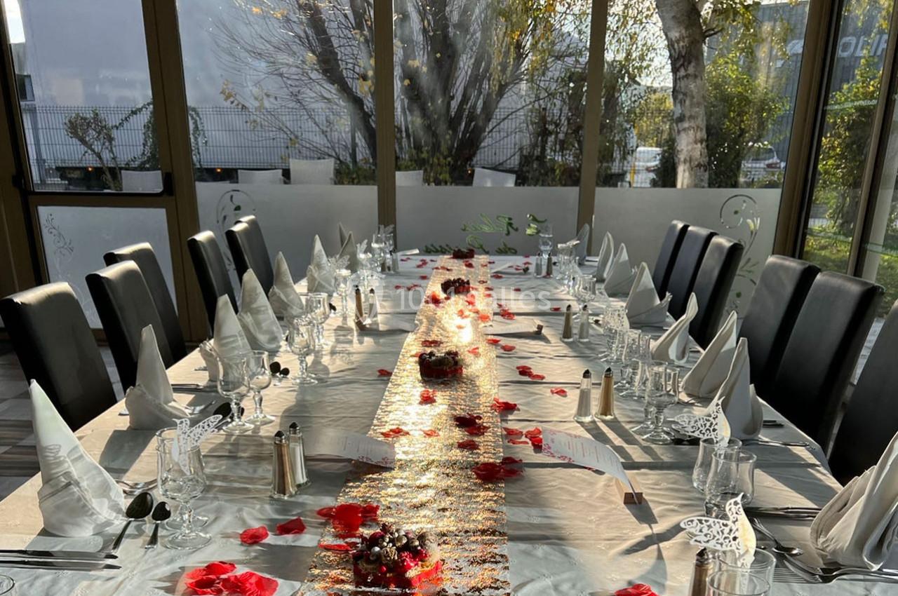 Table dressée avec une nappe blanche, pétales rouges, serviettes pliées et couverts, dans une salle lumineuse.