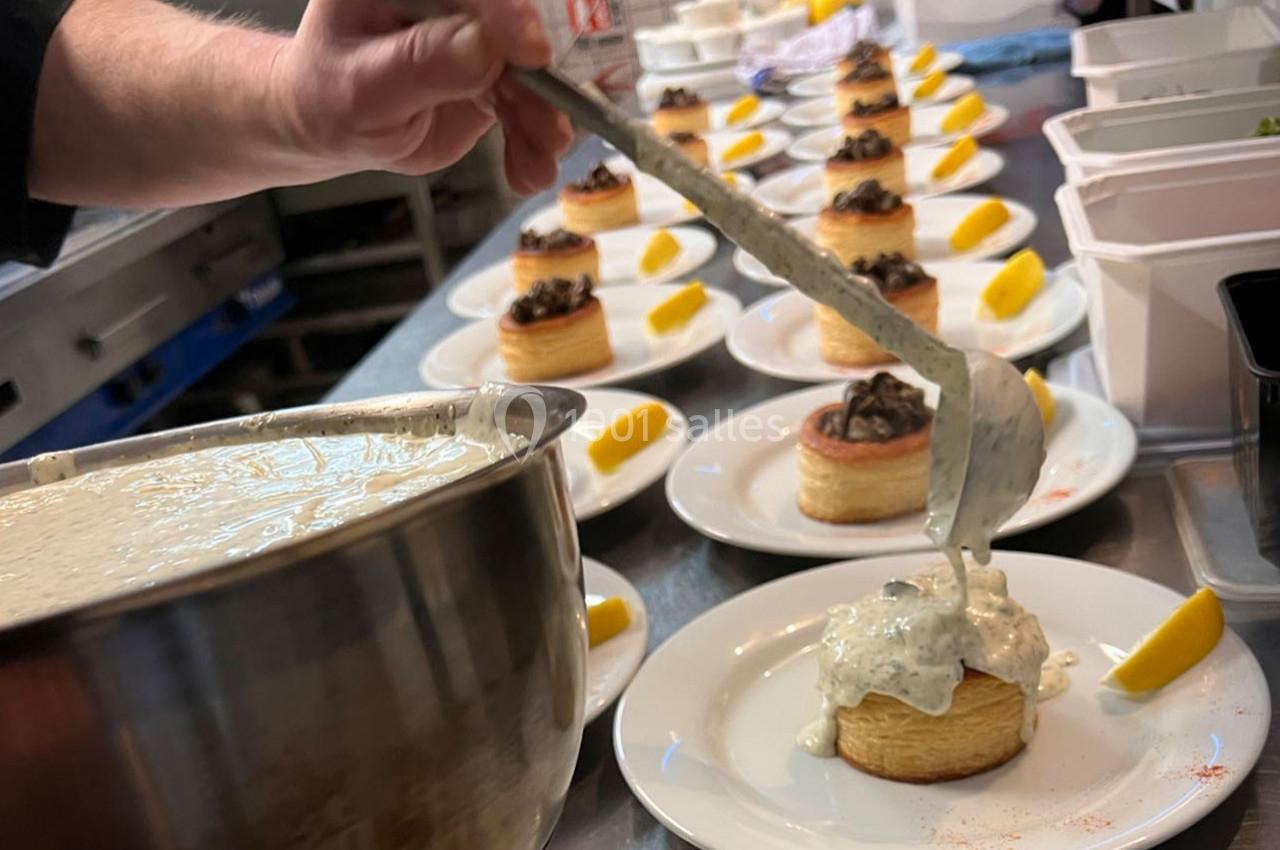 Un chef verse une sauce blanche sur des bouchées garnies, alignées sur des assiettes dans une cuisine professionnelle.