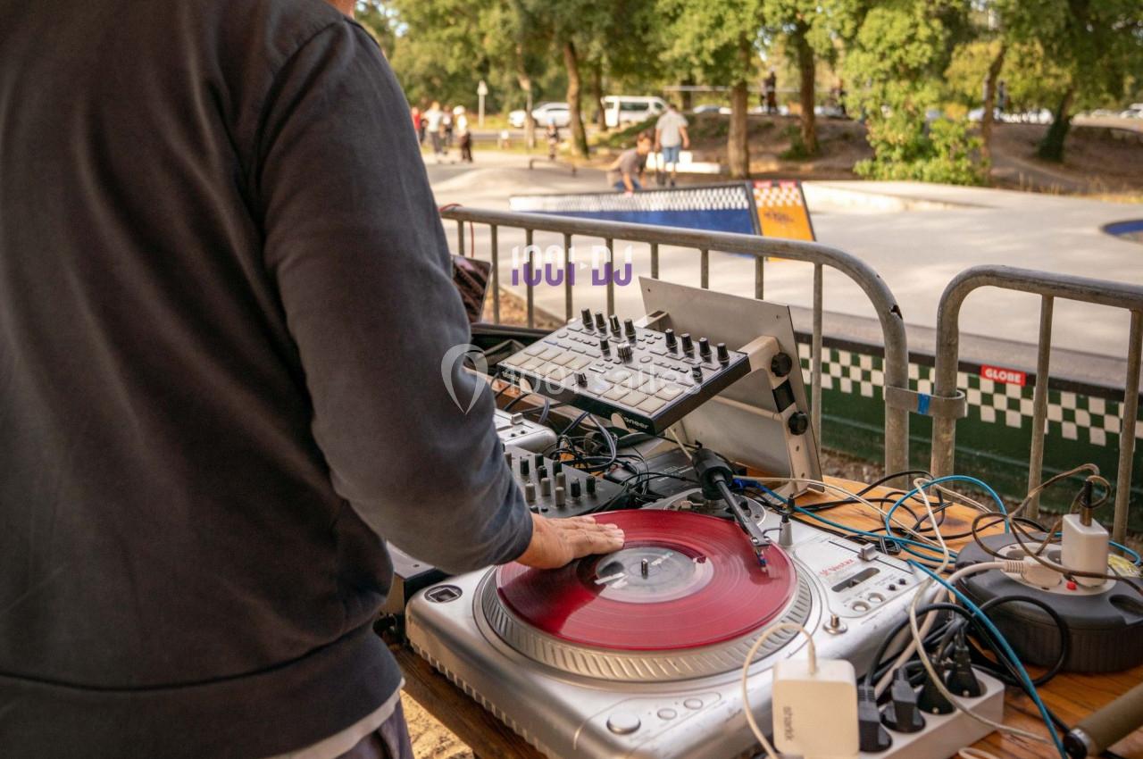 Un DJ manipule un disque vinyle rouge sur une platine, entouré d'équipements audio en extérieur près d'un skatepark.