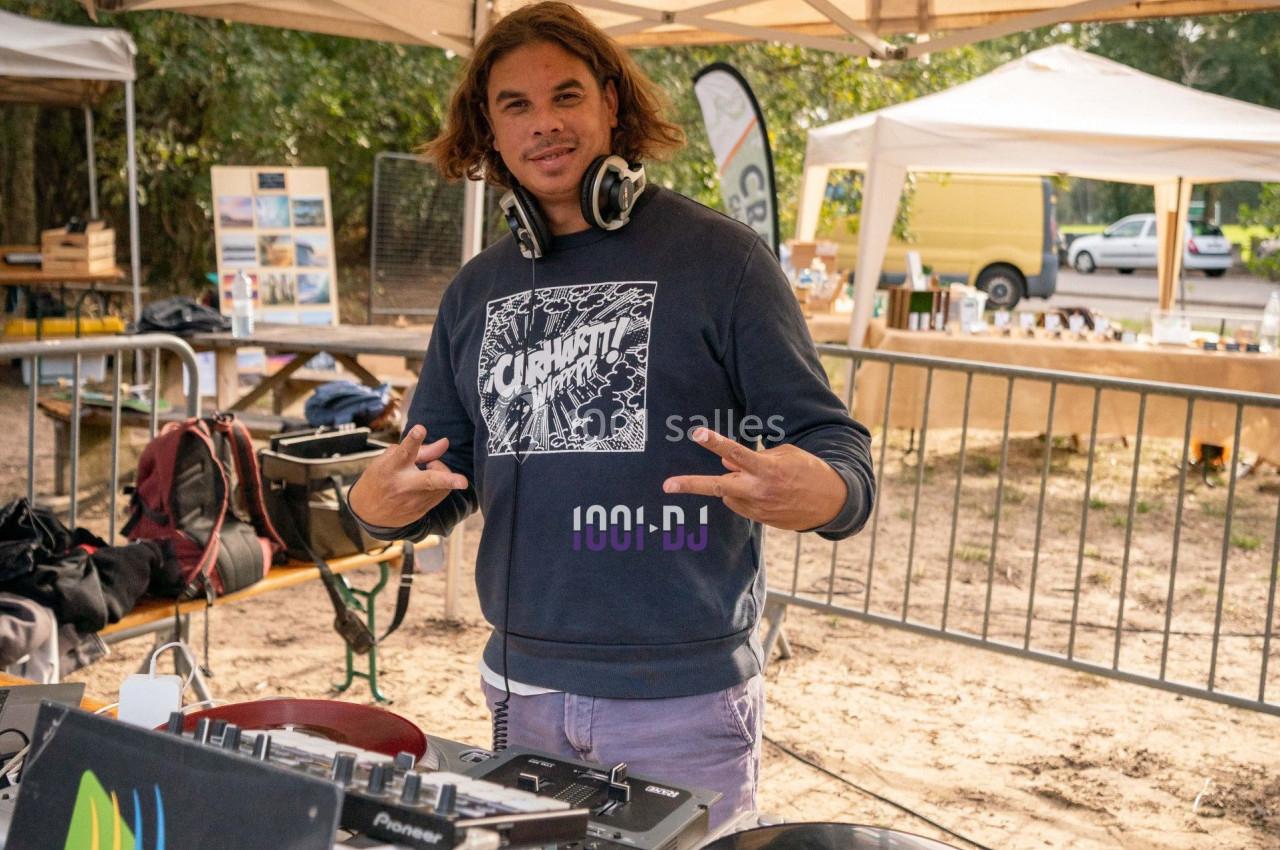 Un DJ souriant devant une table de mixage en plein air, sous une tente lors d'un événement.
