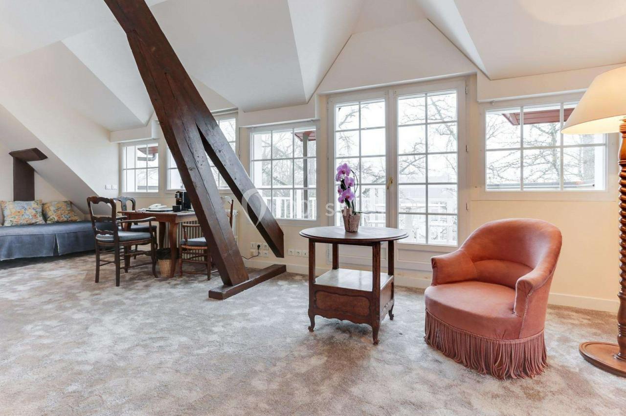 Salon lumineux avec poutre apparente, fauteuil, table basse, bureau et grandes fenêtres donnant sur l'extérieur.