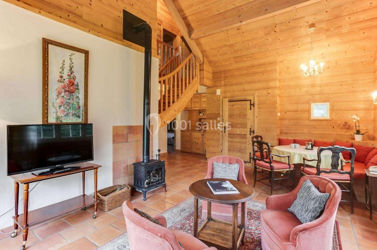 Salon chaleureux avec murs en bois, poêle à bois, télévision, coin repas et escalier menant à l'étage.