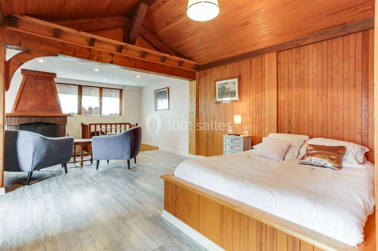 Chambre lumineuse avec lit double, murs en bois, plafond en poutres apparentes, coin salon et cheminée.