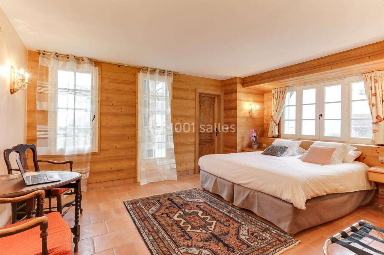 Chambre lumineuse avec murs en bois, lit double, tapis coloré, fenêtres avec rideaux et mobilier en bois.