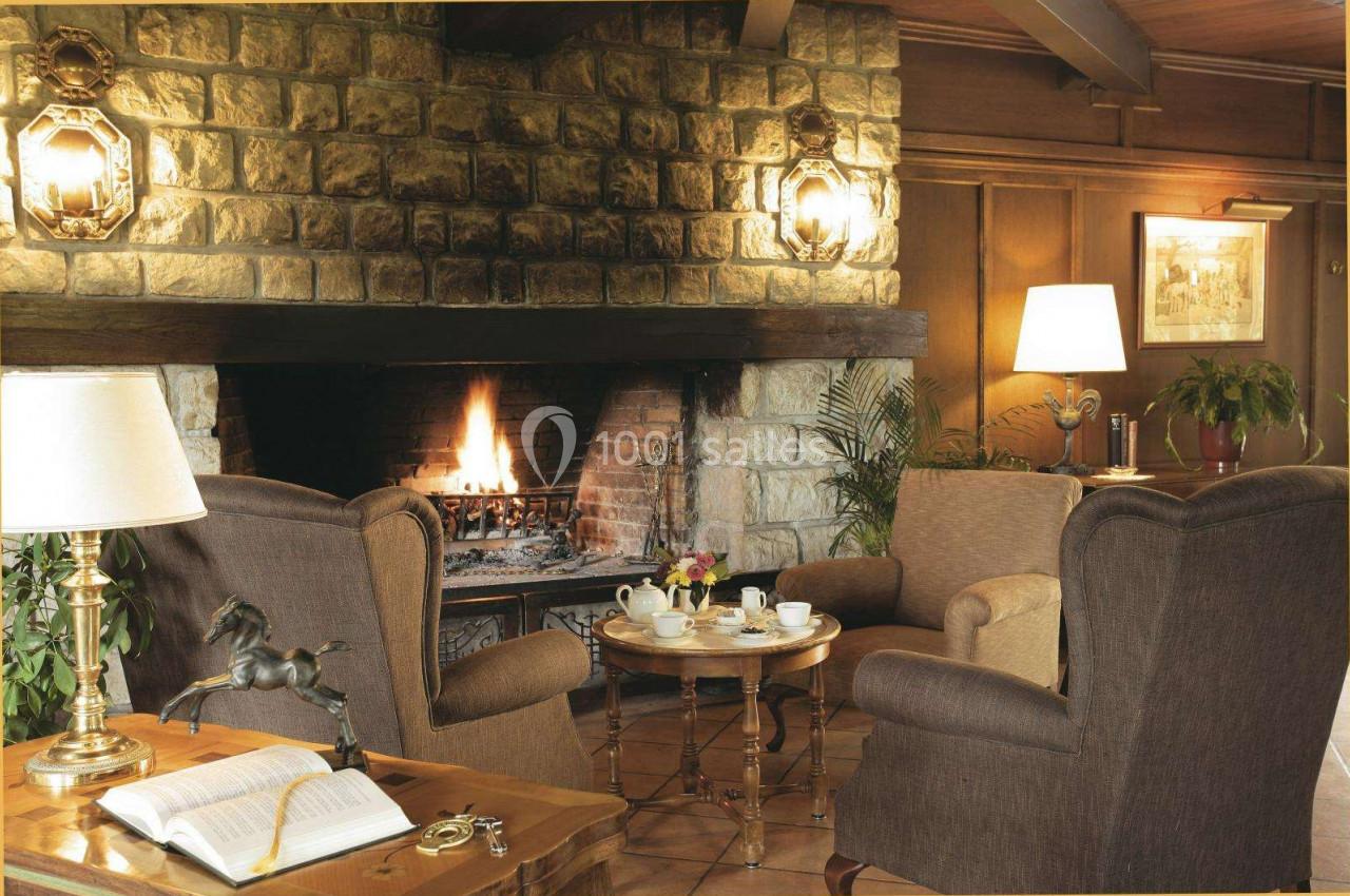 Salon chaleureux avec cheminée en pierre allumée, fauteuils confortables, table basse avec service à thé et lampes allumées.