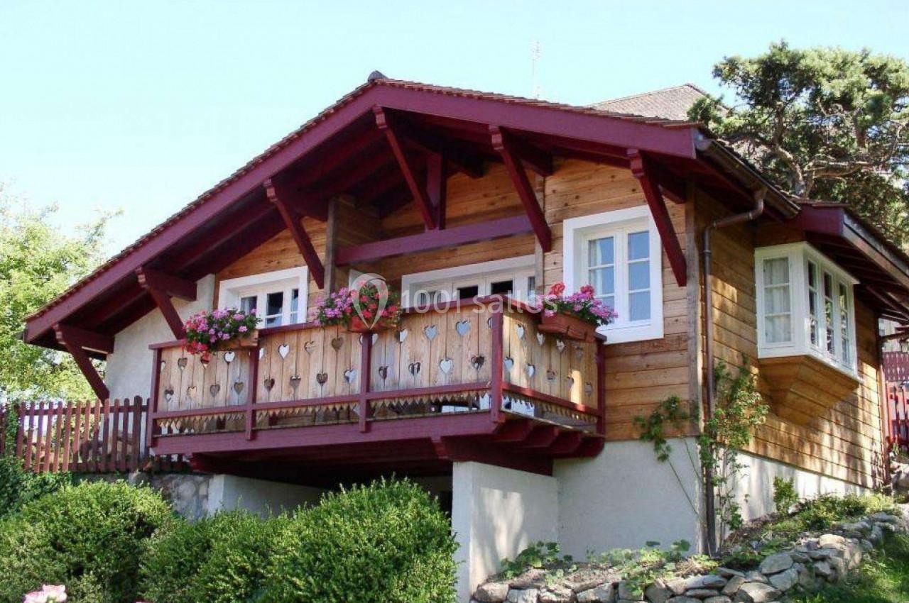 Chalet en bois avec balcon fleuri, entouré de verdure et situé sur un terrain légèrement surélevé.