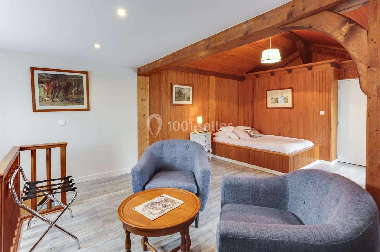 Chambre avec murs en bois, lit double, deux fauteuils gris, table basse et décorations murales.