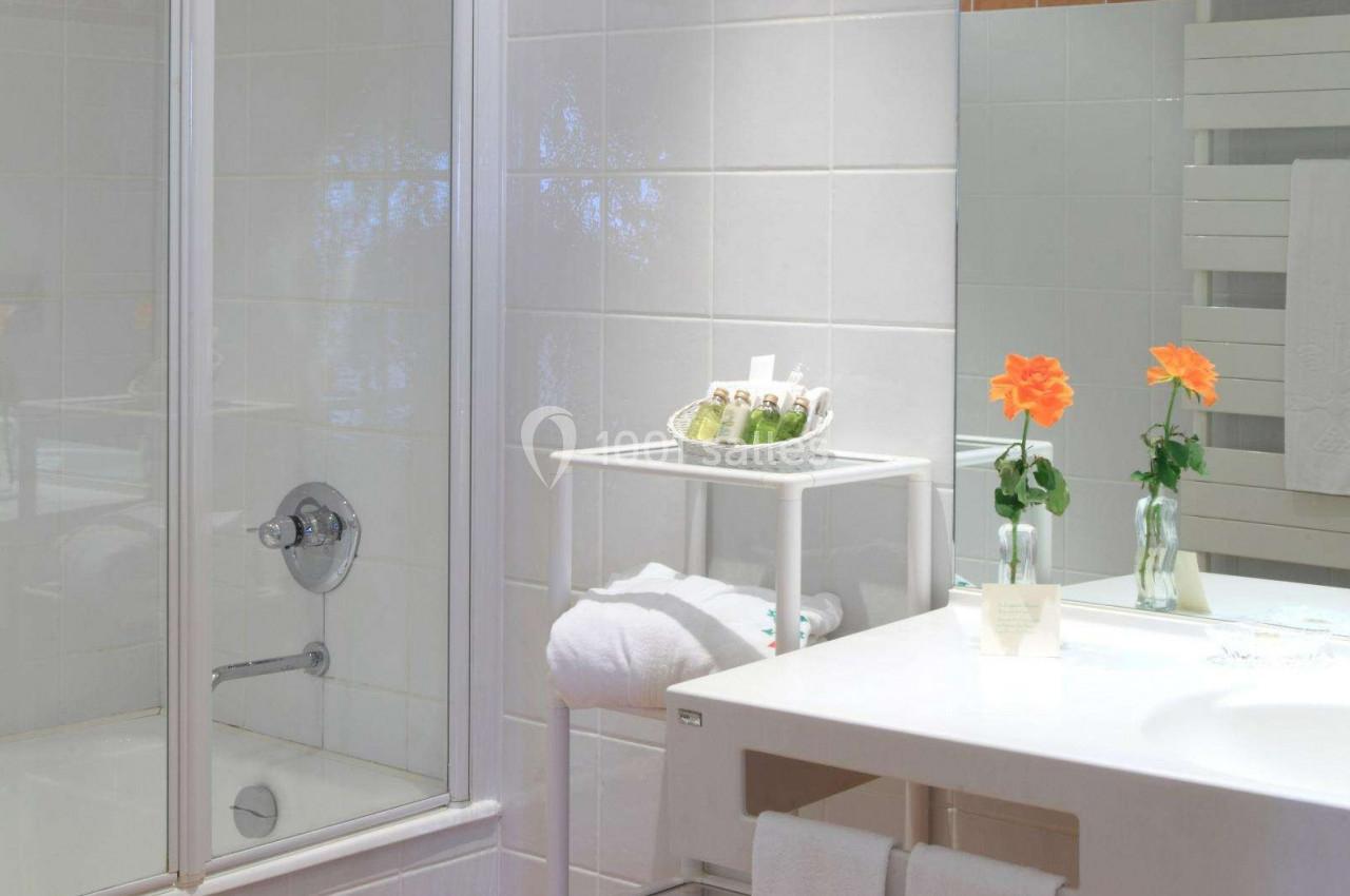 Salle de bain lumineuse avec douche vitrée, lavabo, serviettes pliées, produits de toilette et fleurs décoratives.