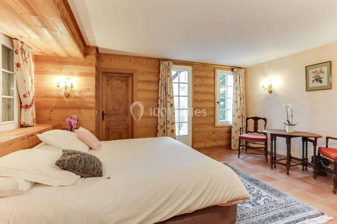 Chambre lumineuse avec murs en bois, lit double, table avec chaises, orchidée et fenêtres donnant sur l'extérieur.
