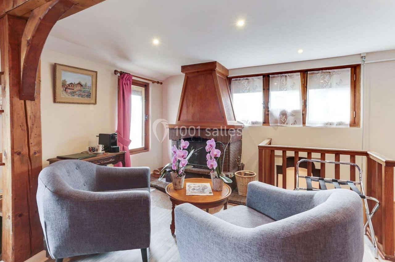 Salon chaleureux avec cheminée en bois, deux fauteuils gris, table basse avec orchidées et fenêtres laissant entrer la…