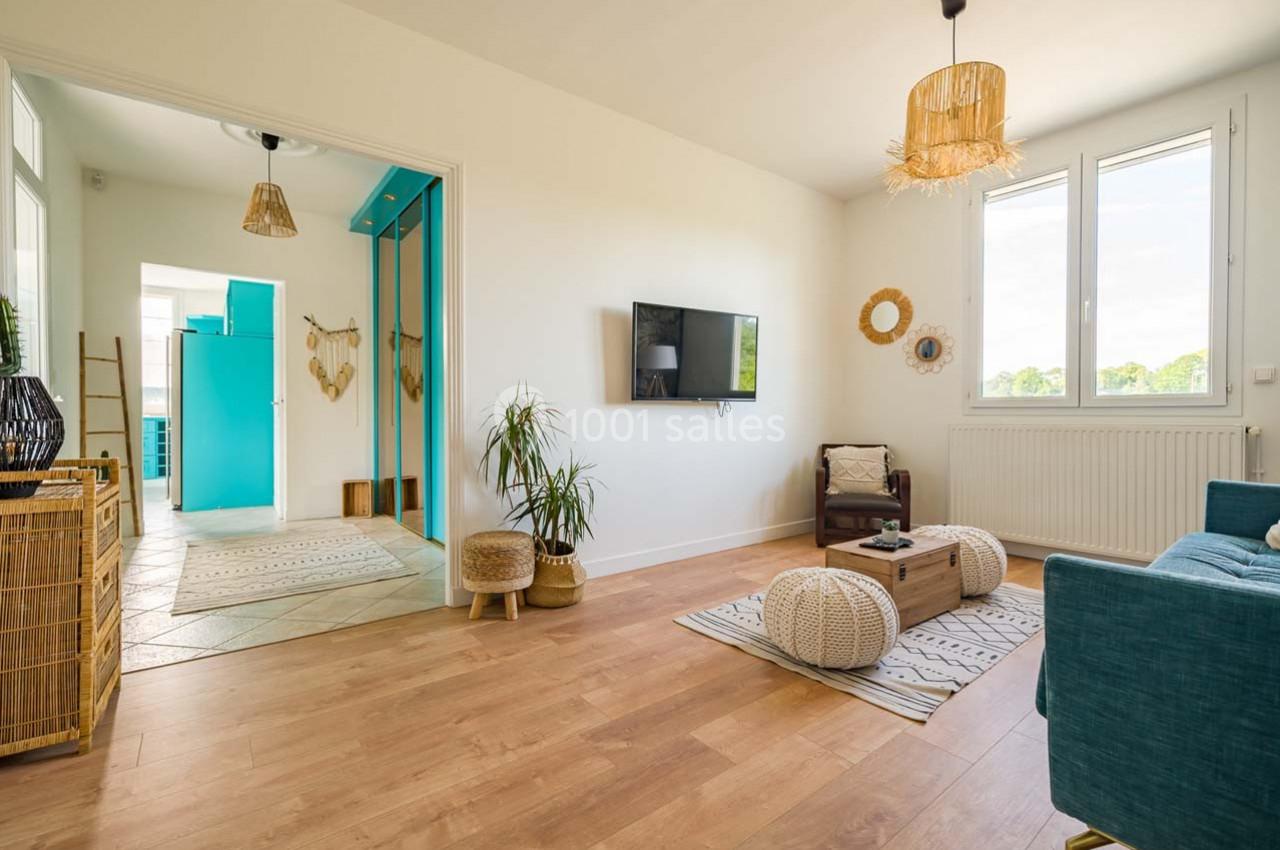 Salon lumineux avec parquet, canapé, table basse, plantes, décoration naturelle et vue sur une cuisine turquoise.