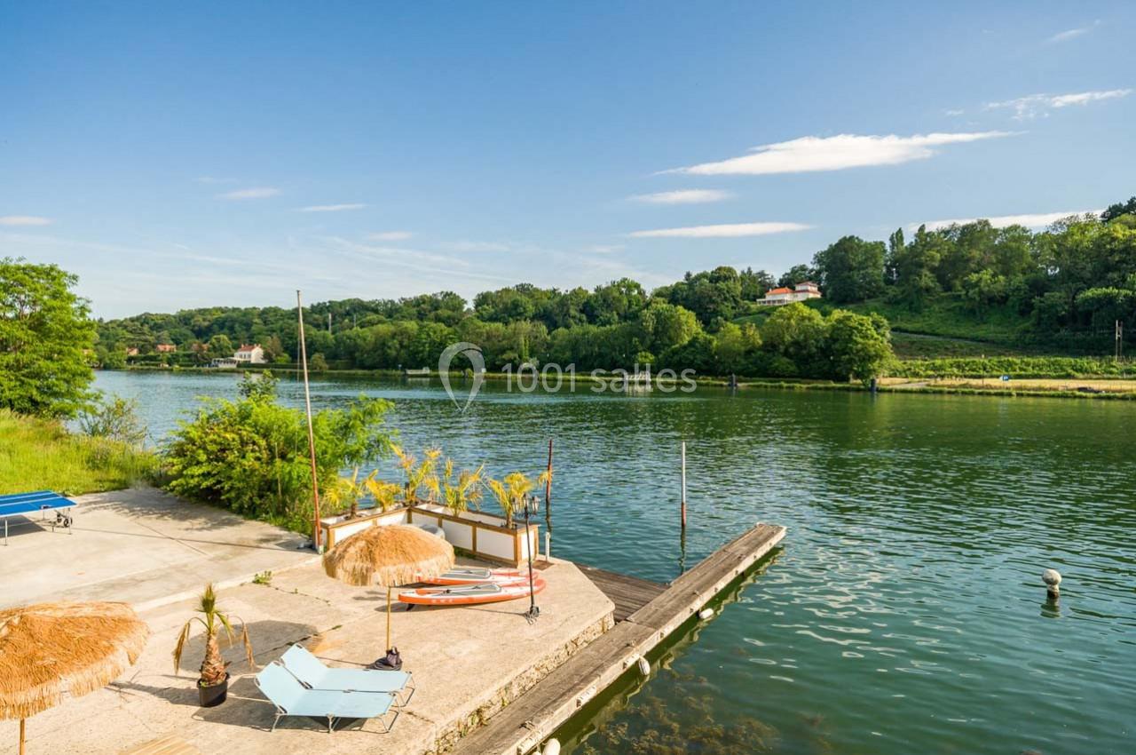 Terrasse avec transats, parasols et ponton en bord de rivière, entourée de verdure sous un ciel dégagé.