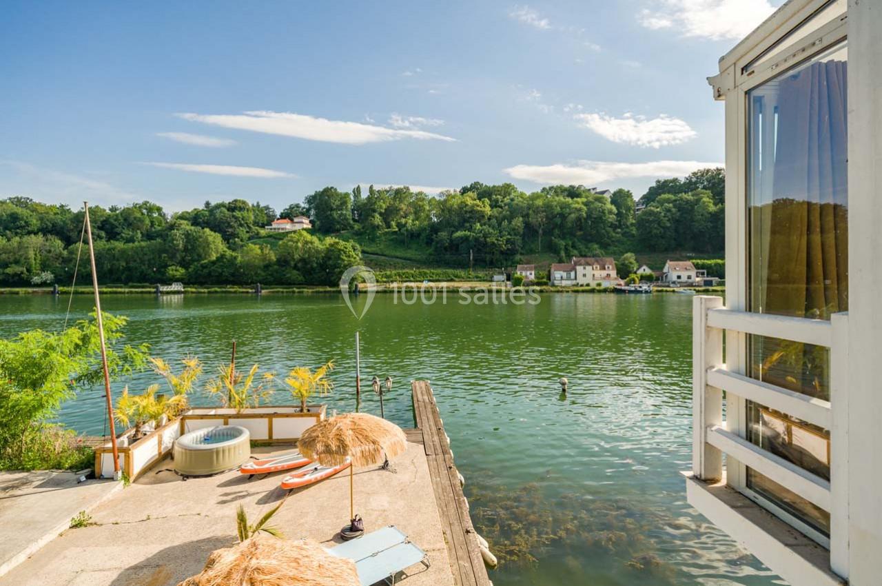 Vue sur une rivière bordée de végétation, avec une terrasse équipée de parasols et un jacuzzi au premier plan.