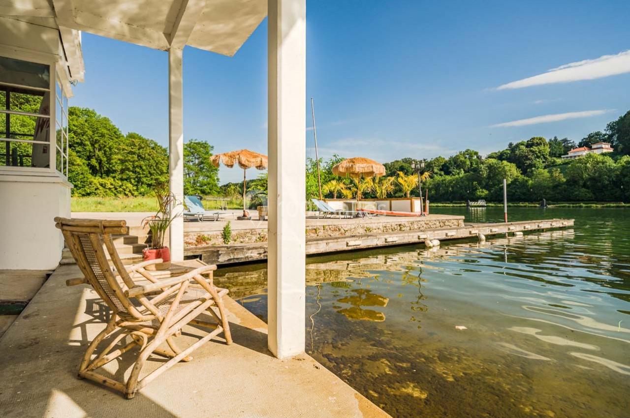 Terrasse au bord d'un lac avec chaise en bois, ponton, parasols en paille et végétation verdoyante en arrière-plan.