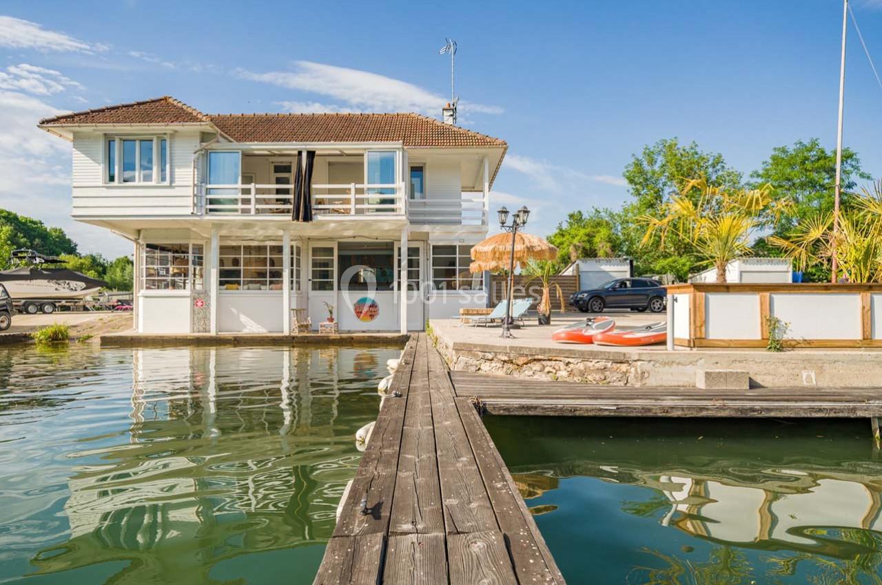 Maison blanche sur pilotis au bord d'un canal, avec terrasse aménagée, kayaks et végétation environnante.