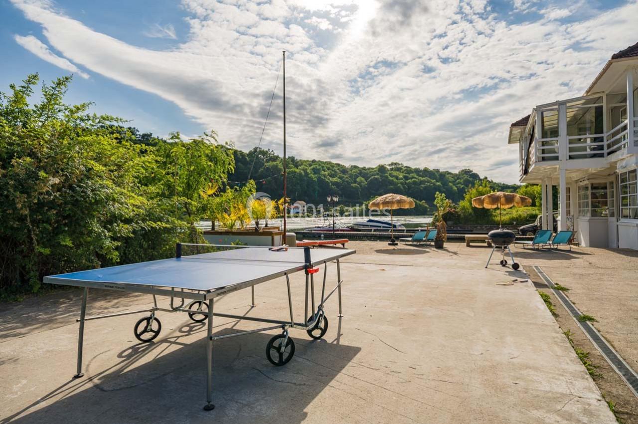 Table de ping-pong sur une terrasse ensoleillée, avec vue sur un lac, des parasols et des collines verdoyantes.