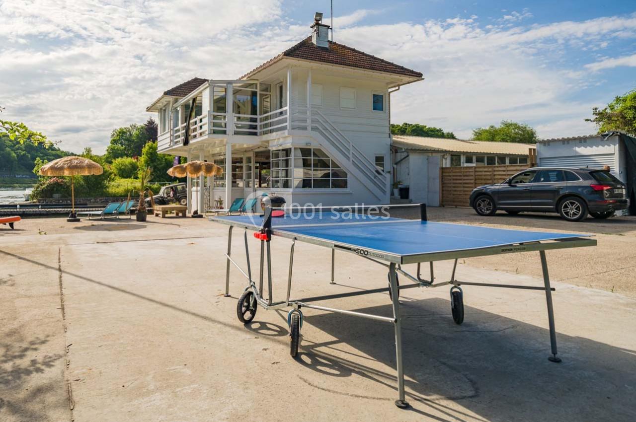 Table de ping-pong sur une terrasse ensoleillée devant une maison blanche à deux étages, avec parasols et voiture garée.