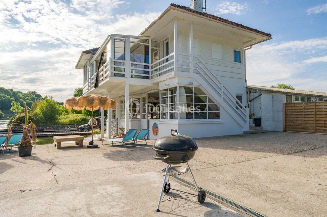 Maison moderne avec terrasse, barbecue au premier plan, transats et parasol près d'une piscine, sous un ciel dégagé.