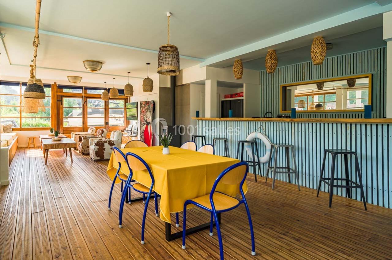 Salle lumineuse avec tables jaunes, chaises bleues, bar en bois et grandes fenêtres donnant sur l'extérieur.